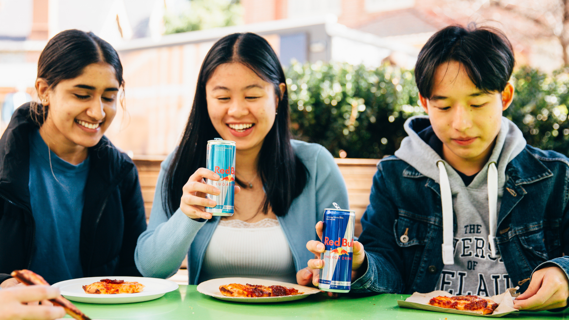 Three students sit at a green table eating pizza on paper plates and drinking red bull.