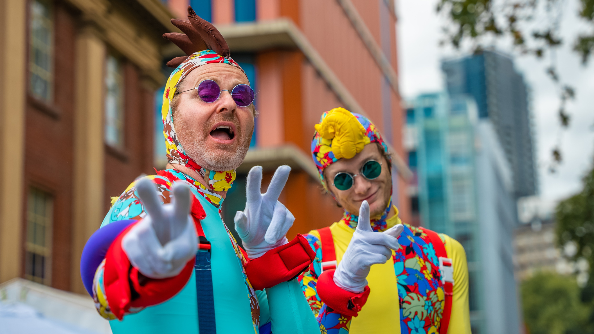 Two performers gesture at the camera in bright blue, green, yellow and red costumes.